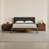Minimalist bedroom with a wooden bed frame, gray upholstered headboard, white bedding, and two matching wooden nightstands. One nightstand has a small plant and bowl, the other a lamp and books. Beige rug and neutral walls.