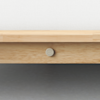 Close-up of a light wood bed frame with a round metal screw in the center engraved with TRUMAN, against a gray floor and a white background.