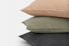 Three stacked pillows with plain covers in different colors: light pink on top, sage green in the middle, and dark gray on the bottom, set against a light background.
