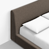 Close-up of a modern bed with a grey upholstered headboard and frame, white pillows, and a white sheet on a mattress, set against a white wall and light grey floor.