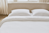 A neatly made bed with a beige upholstered headboard, two white pillows, and a white textured duvet, in a bright room with light curtains and windows in the background.