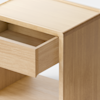 A minimalist wooden cabinet viewed from the side, with a single drawer partially open, standing on a smooth, light-colored floor against a plain white background.