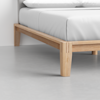 A close-up of the corner of a wooden bed frame with light-colored wood, white bedding, and a grey upholstered headboard, set on a smooth, light grey floor.