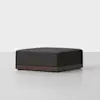 A modern, square, dark gray ottoman with a wooden base sits on a light gray floor against a plain white background.