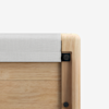 Close-up of the corner of a wooden bed frame with light-colored wood and a light gray fabric covering the top edge, against a plain white background.