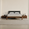 A minimalist bedroom with a wooden bed frame, gray headboard, white bedding, two wooden nightstands, a small tree branch decor on the left, and a lamp on the right, all set on a light gray rug.