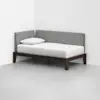 A minimalist daybed with a dark wooden frame, light gray upholstered backrest, and white bedding with a single pillow, placed in the corner of a white room with a smooth floor.