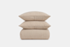 Three beige, textured cushions stacked neatly on top of a matching folded blanket, all with a subtle grid pattern, against a plain white background.
