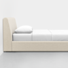 A side view of a minimalist beige upholstered bed with a white pillow and white bedding, placed against a plain white wall on a smooth gray floor.