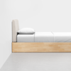 A minimalist wooden bed with a light-colored upholstered headboard, white sheets, a pillow, and a neatly made bed in a clean, modern room with a gray floor and plain white wall.