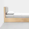 A minimalist wooden bed with a light gray upholstered headboard, a white pillow, and white bedding, placed against a white wall on a gray floor. The bed is shown from the side.