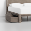 A wooden bed frame with white bedding, featuring a large under-bed storage drawer that is pulled out. The background is minimal with a plain white wall and light gray floor.