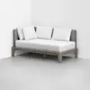 A modern daybed with a light wood frame, gray upholstered backrest, and three white pillows, placed in the corner of a minimalist white room.