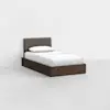A single bed with a dark wood frame and an upholstered, grey headboard. The bed is made with white sheets and a pillow, placed in an empty, minimalist room with white walls and floor.