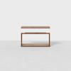 A minimalist wooden side table with a rectangular top, open sides, and a flat base, placed on a light gray floor against a plain white background.