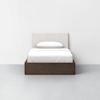 A neatly made twin bed with white bedding, a white pillow, a light upholstered headboard, and a dark wooden frame, set against a plain white wall and gray floor.