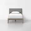 A single bed with a light wooden frame and a gray upholstered headboard, dressed in white bedding with a single white pillow, set against a plain white wall and a smooth light gray floor.