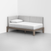 A minimalist daybed with light wood frame, white mattress, single white pillow, and light gray upholstered backrests, positioned in the corner of a plain white room with a smooth gray floor.