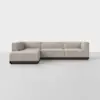 A modern L-shaped sectional sofa with light beige upholstery and a dark wooden base, positioned against a plain white background and floor.