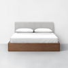 A modern bed with a light gray upholstered headboard, white bedding, two white pillows, and a wooden platform frame, set against a plain white wall and a light gray floor.