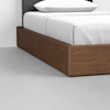 A close-up view of a bed with a wooden platform frame, white bedding, and a white pillow, situated on a smooth, light gray floor.
