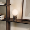 A glowing spherical white lamp with a dark brown marble cylindrical base sits on a dark wood shelf, beside a framed artwork, with books and a bowl visible on nearby shelves.