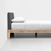 A minimalist bed with a wooden frame, gray upholstered headboard, and white bedding and pillow, set against a plain white wall and floor.