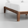 A close-up view of a wooden bed frame with clean lines and a modern design, featuring white bedding and pillows in a minimalistic room with a light gray floor.