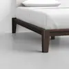 A close-up of a modern bed with a dark wooden frame and white bedding, focusing on the corner and legs of the bed against a light-colored floor and wall.