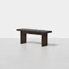 A minimalist wooden bench with a dark brown finish and a black cushioned seat, positioned on a light-colored floor against a plain white background.