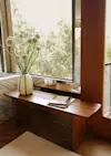 A wooden bench sits by a large window with trees outside. On the bench is a vase with white allium flowers and a stack of books. A mirror behind the bench reflects the scene. Marble accents are visible nearby.
