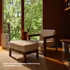 A modern lounge chair with matching ottoman and a small side table, all in walnut wood and ivory fabric, sit in a sunlit room with large windows overlooking green trees. Text in the corner describes the furniture.