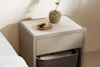 A modern beige nightstand with a stone top holds a small ceramic vase with a few flowers and a round tray with decorative objects. Below, a shelf contains a soft gray storage basket with a white cloth inside.