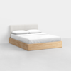 A modern bed with a light wood platform base, white upholstered headboard, and neatly made with white bedding and two pillows, set in a minimalist room with white walls and floor.