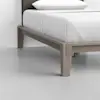 A close-up view of the corner of a modern wooden bed frame with clean lines, featuring white bedding and a pillow, set on a smooth, light-colored floor against a plain wall.