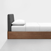 A minimalist bed with a wooden frame, a dark gray upholstered headboard, and white bedding, pillow, and sheets, set against a plain white wall and gray floor.