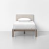 A single bed with a beige upholstered headboard, white bedding, and a pillow, set against a plain white wall in a minimalist room with a light gray floor.