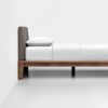A minimalist wooden bed frame with a padded gray headboard, dressed in white bedding and a white pillow, positioned against a plain white wall and floor.