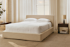 A modern bedroom with a beige upholstered bed frame, white bedding, two pillows, a bedside table with a lamp, a plant, and large windows with sheer curtains letting in natural light.