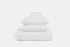 A neatly stacked set of white bedding, including folded sheets, pillowcases, two pillows, and a comforter, arranged in a pyramid shape on a white background.