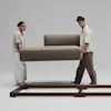 A woman and a man lift a cushioned sofa section together, preparing to place it onto a wooden frame in a minimal, light-colored studio setting.
