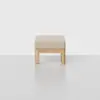 A minimalist ottoman with a light wood frame and a beige cushioned top, placed on a smooth, light-colored floor against a plain white background.