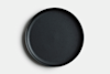 A round, matte black ceramic plate with a slightly raised edge, photographed from above on a white background. The plate is empty and has a simple, modern design.