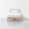A neatly made single bed with a light-colored upholstered headboard, white bedding, and a light wood frame, placed against a plain white wall on a light gray floor.