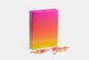 A rectangular box with a pink-to-yellow gradient stands upright on a white surface. Several colorful puzzle pieces are scattered on the ground in front of the box.