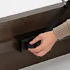A person adjusts a black metal lever attached to a dark wooden beam, turning a round knob with their right hand. The persons sleeve is beige.