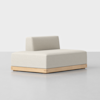 A modern, minimalist beige lounge chair with a low backrest and wooden base, placed on a light gray floor against a plain white background.