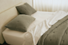 A neatly made bed with a beige upholstered headboard, white sheets, and a textured gray comforter and matching pillows. Soft natural light filters through sheer curtains in the background.