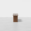 A minimalist wooden bench with a flat seat and solid rectangular base, placed on a light gray floor against a plain white background, viewed from the side.