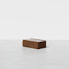 A small, rectangular wooden box with a lid sits on a smooth, light grey surface against a plain white background.